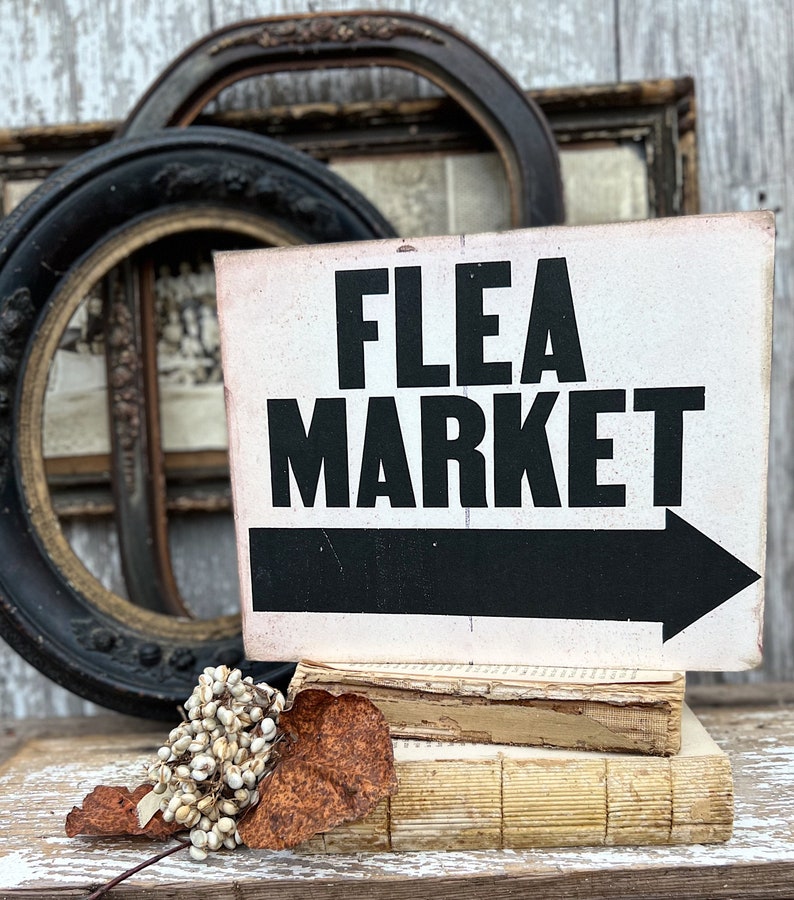 May include: A white sign with black text that reads 'Flea Market' with a black arrow pointing to the right. The sign is propped up on three old books.
